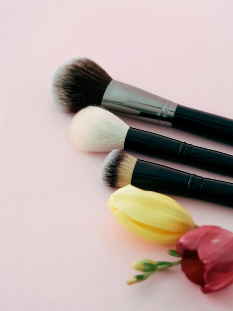 Artistic composition of makeup brushes and flowers against a soft pink backdrop.